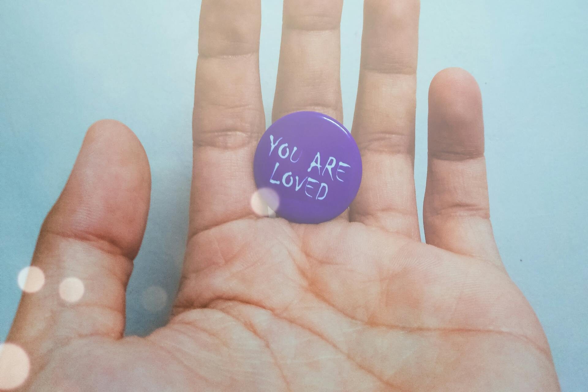 Close-up of a hand holding a purple pin with 'You Are Loved' message on a bright background.